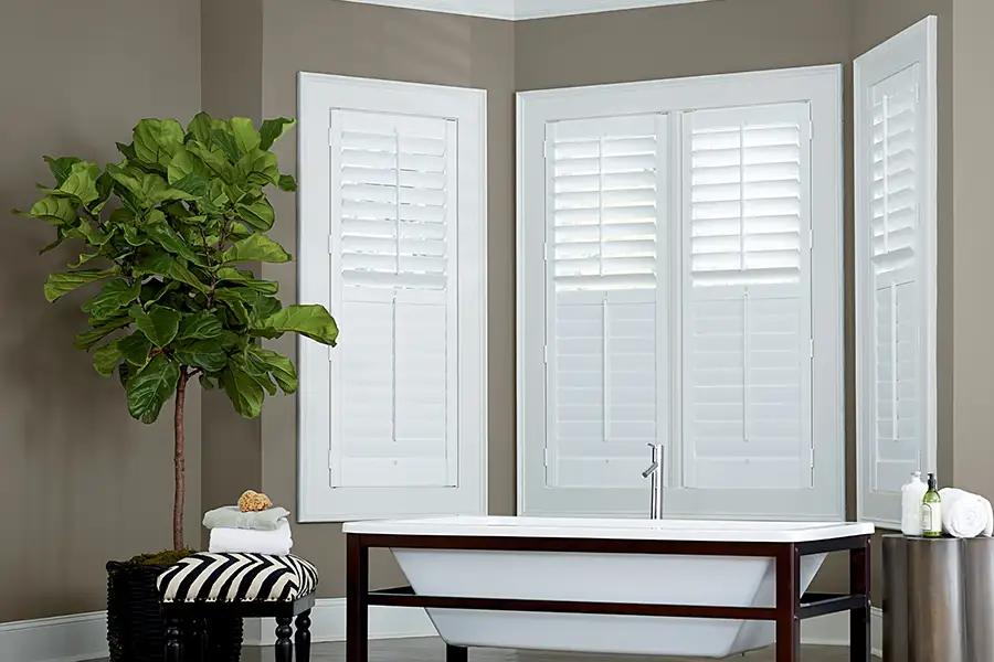For a large bay window, choose energy-efficient Polywood® shutters that will help regulate room temperature. White Polywood shutters on a bay window in a brownish gray wall bathroom