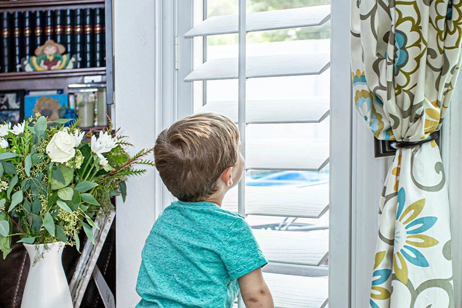Plantation shutter louvers are simple to adjust, even for a small child. Boy looking through a plantation shutter with tilted louvers