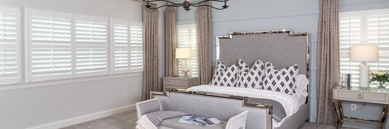 A blue and gray bedroom with elegant furniture, curtains and plantation shutters, and chandelier light