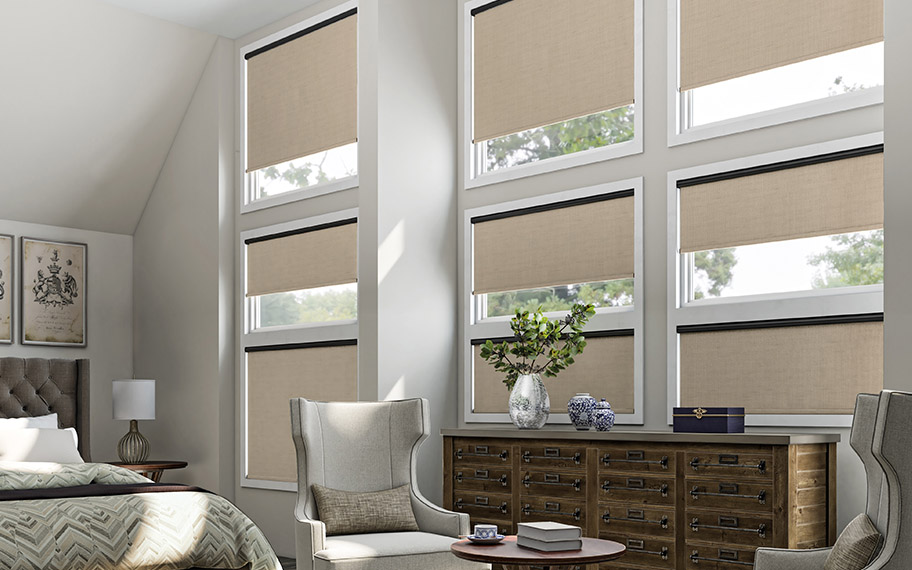 Motorized shades are the perfect solution for hard-to-reach windows. Beige motorized roller shades in a large bedroom with floor-to-ceiling windows.