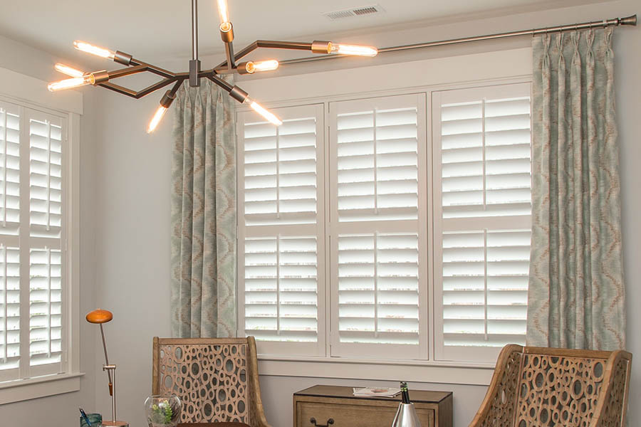 Insulate your windows and add eye-catching style when you pair interior shutters with curtains. sage green and brown wavy pattern curtains with white polywood shutters in an office
