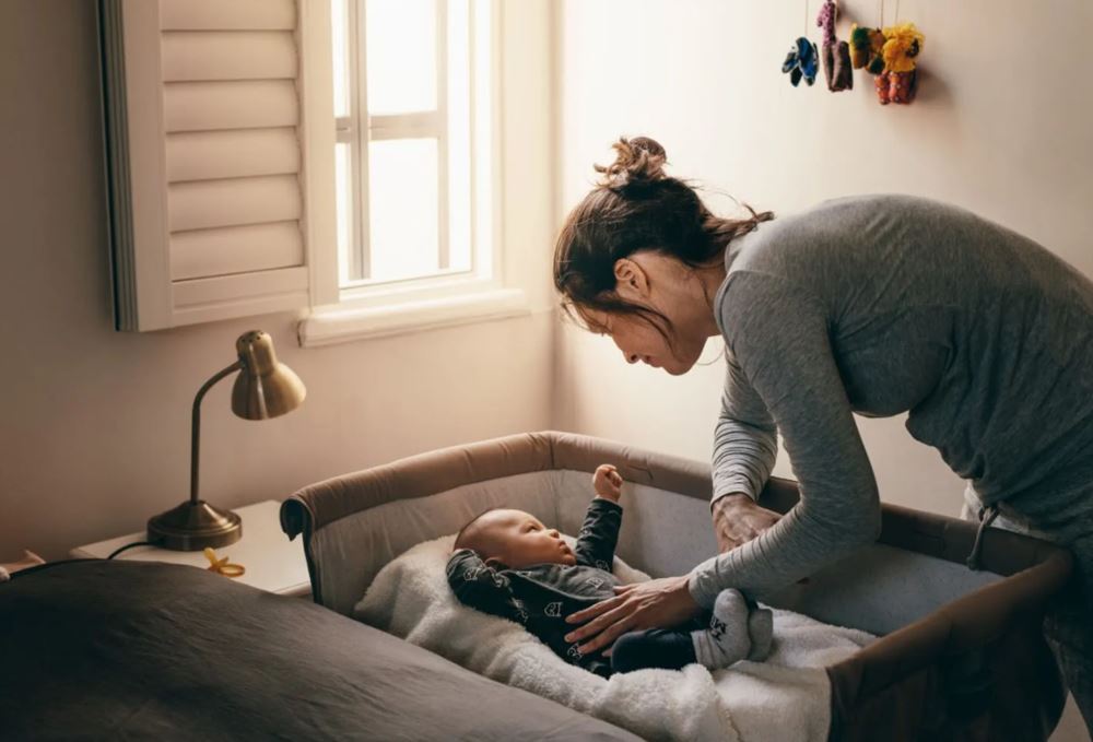 Darkness, privacy, and energy efficiency--Polywood shutters provide everything your little one needs for good sleep. A mother looking at her baby in its bassinet next to a window with Polywood shutters.